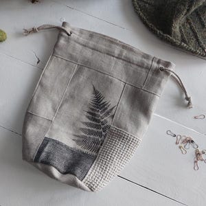 Może przedstawiać: Beżowa torba na projekty ze sznurkiem i nadrukiem paproci. Torba wykonana jest z różnych paneli materiałowych, w tym z teksturowanego splotu waflowego i czarno-białego geometrycznego wzoru. Przeznaczona do projektów dziewiarskich lub szydełkowych.