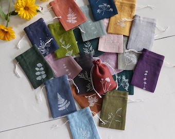 May include: A collection of small, colorful drawstring bags made of linen. Each bag features a white botanical print, such as ferns and wildflowers. The bags are arranged on a white wooden surface, with yellow flowers in the background.