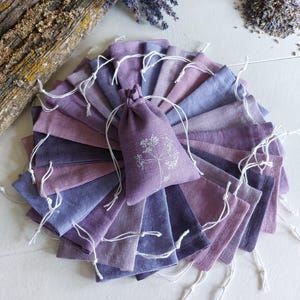 Peut inclure: Un éventail de petits sachets en lin violets, chacun avec un cordon de serrage. Un sachet central avec un motif floral blanc est présenté. De la lavande séchée et un morceau de bois sont en arrière-plan, suggérant un thème naturel.