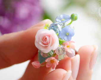 May include: A delicate floral brooch featuring a pink rose and blue forget-me-nots, accented with clear and green beads. The brooch is held in a hand against a blurred background.