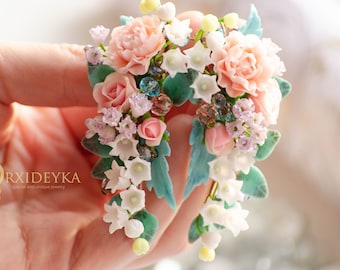 May include: A pair of floral earrings featuring sculpted peach roses, white lily of the valley blossoms, and small light purple flowers. The design includes teal green leaves and translucent blue beads, with the text ORXIDEYKA printed on the side.