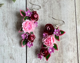 Peut inclure: Une paire de boucles d'oreilles pendantes composées de grappes de fleurs sculptées dans des tons de bordeaux profond, rose clair et violet, agrémentées de feuilles vertes. Les compositions florales sont fixées à des crochets en métal argenté.