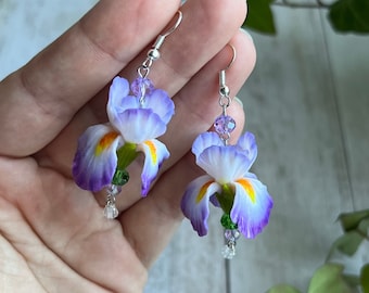 May include: A pair of dangle earrings featuring purple and white iris flowers with yellow centers and green leaves. The earrings are accented with clear and purple crystal beads.