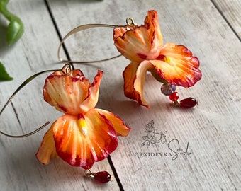 May include: A pair of gold hoop earrings with orange and red flower-shaped charms. The flowers have a yellow center and red edges. The earrings are accented with small red and purple beads.