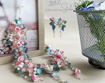 May include: A floral jewelry set featuring a necklace and earrings. The necklace and earrings are adorned with pink, blue, and white flowers, pearls, and silver accents. The set is displayed near a framed picture and a metal mesh container.