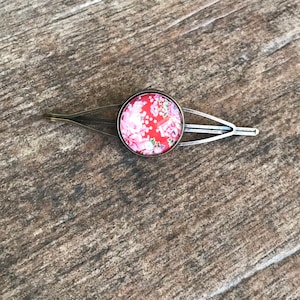 May include: A bronze hair clip with a red and white floral design. The clip is about 2 inches long and has a rounded shape.