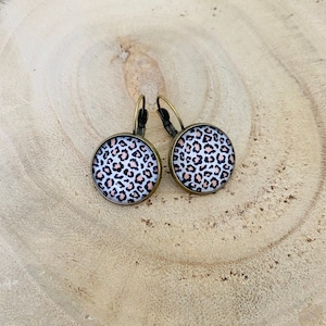 Puede incluir: Un par de pendientes redondos con diseño de estampado de leopardo. Los pendientes tienen un marco de metal color bronce y un fondo blanco con manchas negras y melocotón. Un accesorio elegante.