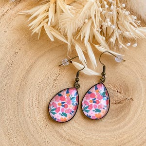Op de afbeelding: Een paar druppelvormige oorbellen met een levendig bloemenpatroon van roze, oranje en blauwgroene bloesems op een lichte achtergrond. De oorbellen zijn gezet in donkerbronskleurige metalen monturen en zijn voorzien van bijpassende bronzen oorhaken.