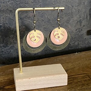 Könnte beinhalten: Ein Paar goldene und rosafarbene Ohrringe mit einem tropischen Blattdesign. Die Ohrringe hängen an einem goldenen Ständer.
