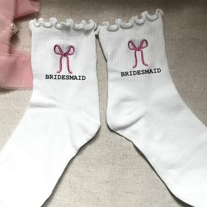 May include: A pair of white ankle socks with ruffled cuffs, each featuring an embroidered pink bow and the black text BRIDESMAID centered below the bow. The socks are made of a ribbed fabric and are displayed side by side.