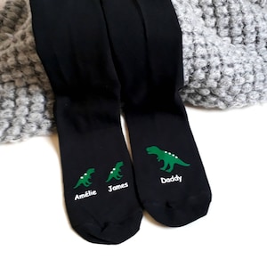 May include: A pair of black socks with green dinosaur graphics. The socks are personalised with the names "Amélie", "James", and "Daddy".