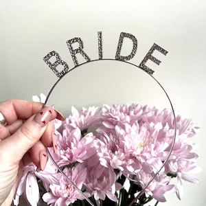May include: A silver-toned headband with the word "BRIDE" spelled out in sparkling, rhinestone-like letters. The headband is held in front of a bouquet of light pink chrysanthemums, creating a celebratory and feminine aesthetic.