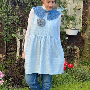 May include: A woman wearing a blue and white gingham dress with a blue crocheted collar and a large gray flower brooch. She is wearing blue jeans and white shoes.