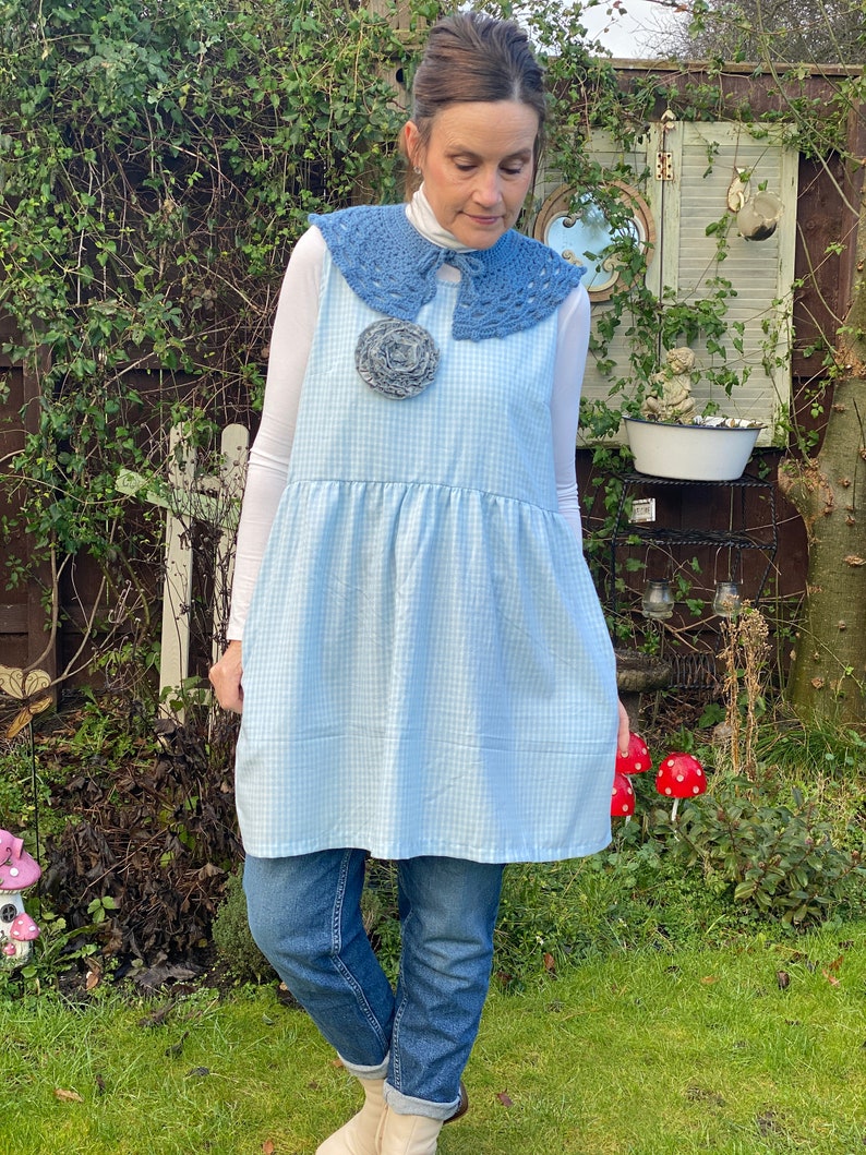 May include: A woman wearing a blue and white gingham dress with a blue crocheted collar and a large gray flower brooch. She is wearing blue jeans and white shoes.