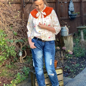 May include: A woman wearing a white blouse with a floral print and a crocheted orange collar. She is also wearing blue jeans with distressed details and white ankle boots.