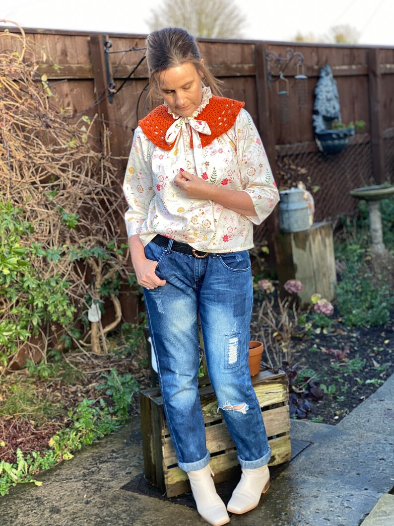 May include: A woman wearing a white blouse with a floral print and a crocheted orange collar. She is also wearing blue jeans with distressed details and white ankle boots.