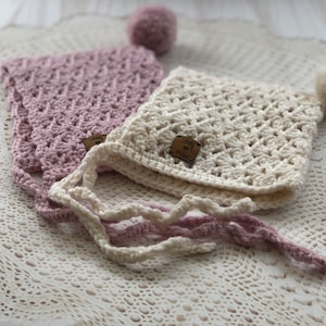 May include: Two crocheted baby bonnets with ties, one in white and one in pink, with a small pom-pom on top of each. The bonnets have a label that says "Loopy & Mabel".