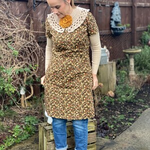 May include: A woman wearing a brown floral dress with a white crochet collar and a yellow flower brooch. She is also wearing blue jeans and white ankle boots.