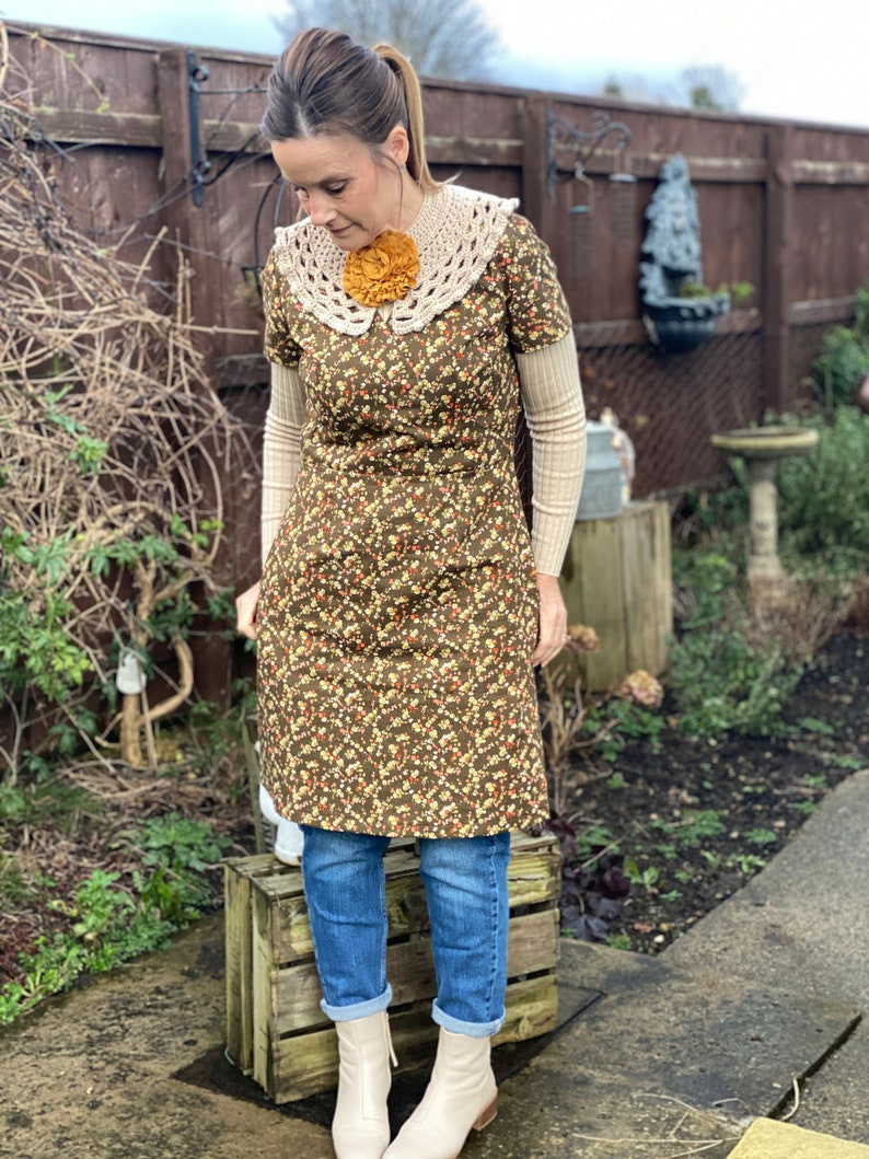 May include: A woman wearing a brown floral dress with a white crochet collar and a yellow flower brooch. She is also wearing blue jeans and white ankle boots.