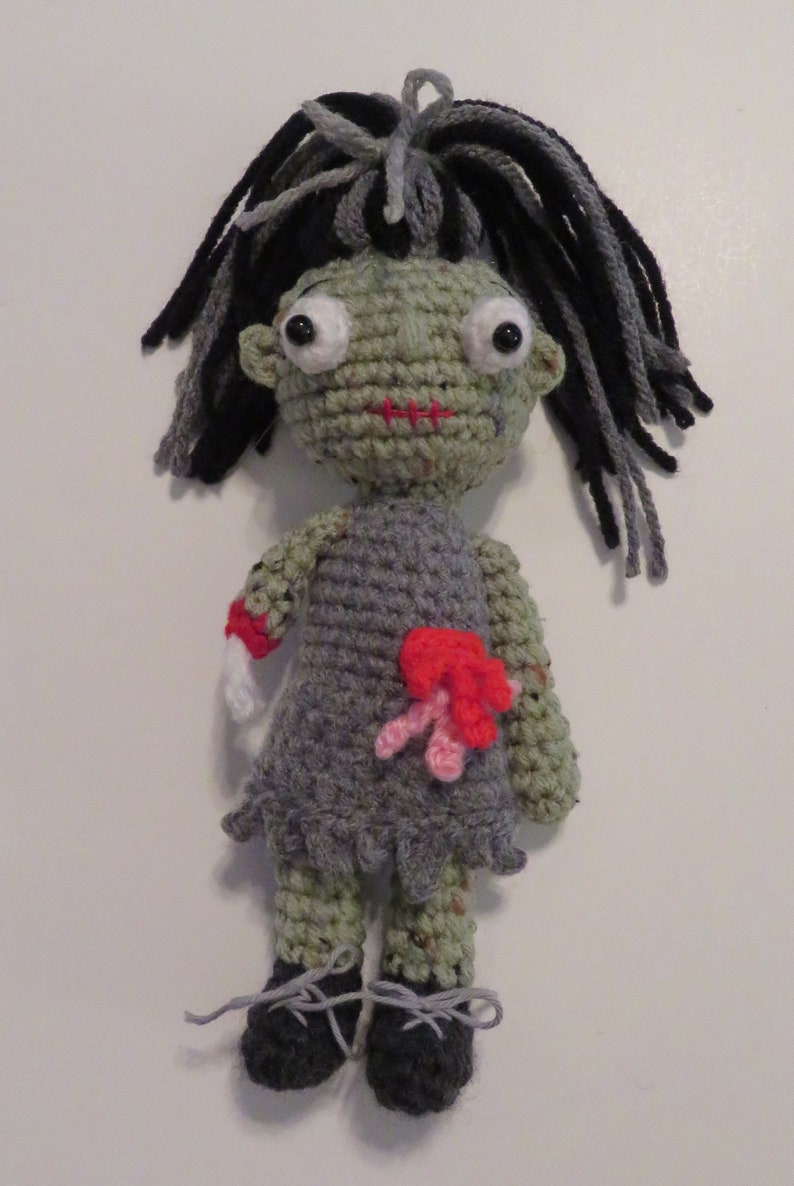 crochet girl zombie with green skin, grey dress and black and grey hair. She has bulging eyes, red scar for mouth, a severed arm with a crochet bone sticking out. She has red and pink intestines coming out of her belly, a red scar one one arm and leg