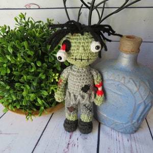 Crochet boy zombie with green skin, grey clothing, a dislocated eye and a partially severed arm that shows a white bone between the upper and lower arm and red yarn for blood. He has black messy hair, an embroidered scar for a mouth.