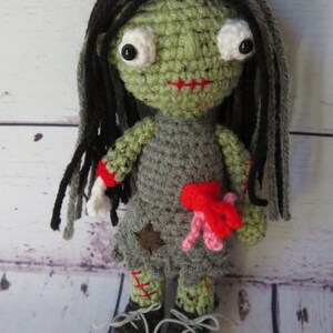 crochet girl zombie with green skin, grey dress and black and grey hair. She has bulging eyes, red scar for mouth, a severed arm with a crochet bone sticking out. She has red and pink intestines coming out of her belly, a red scar one one arm and leg