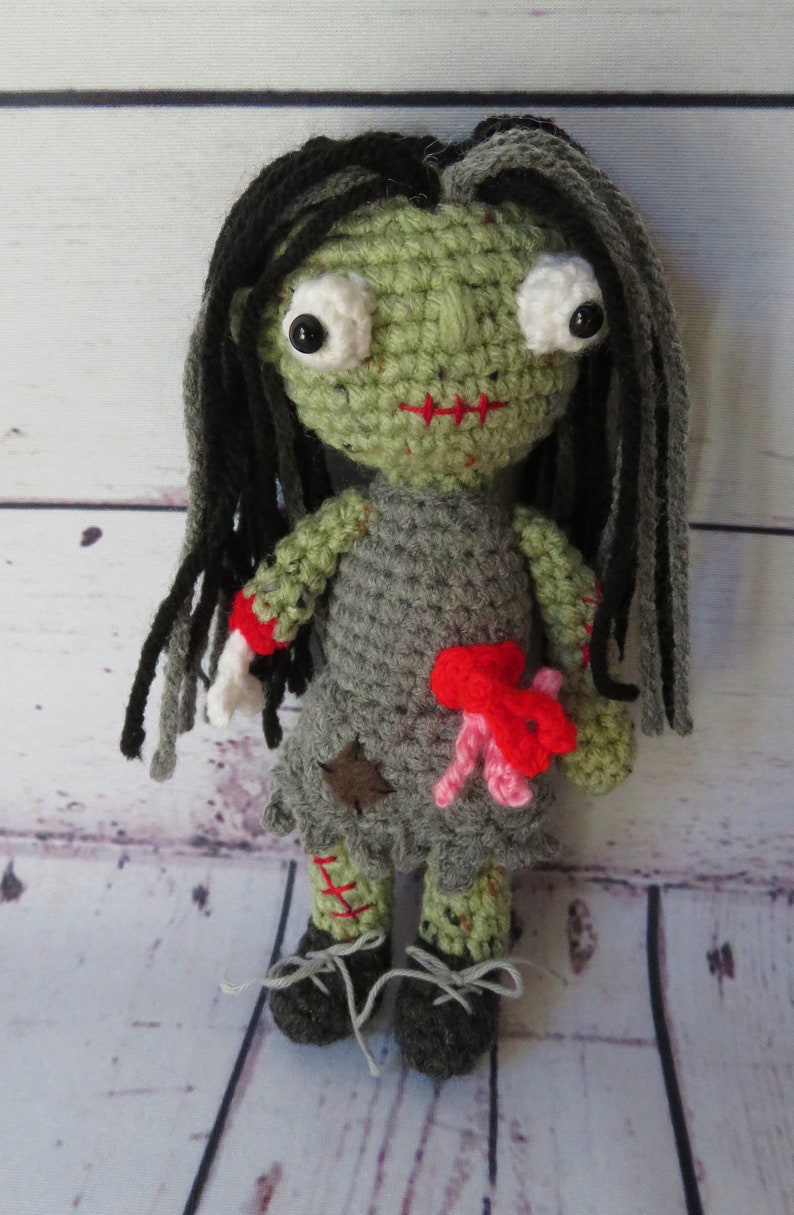 crochet girl zombie with green skin, grey dress and black and grey hair. She has bulging eyes, red scar for mouth, a severed arm with a crochet bone sticking out. She has red and pink intestines coming out of her belly, a red scar one one arm and leg