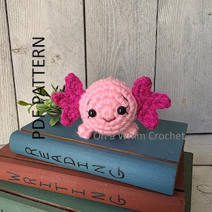 Op de afbeelding: Een handgemaakte roze gehaakte axolotl met felroze kieuwen en zwarte veiligheidsoogjes. De axolotl zit op een stapel boeken met de woorden "Reading" en "Writing" zichtbaar. De tekst "PDF PATTERN" staat aan de linkerkant.