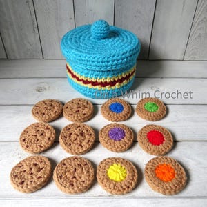 May include: A crocheted cookie jar and cookies. The jar is turquoise with a red and yellow stripe. The cookies are brown with colorful centers. The text "Gina Whim Crochet" is visible.