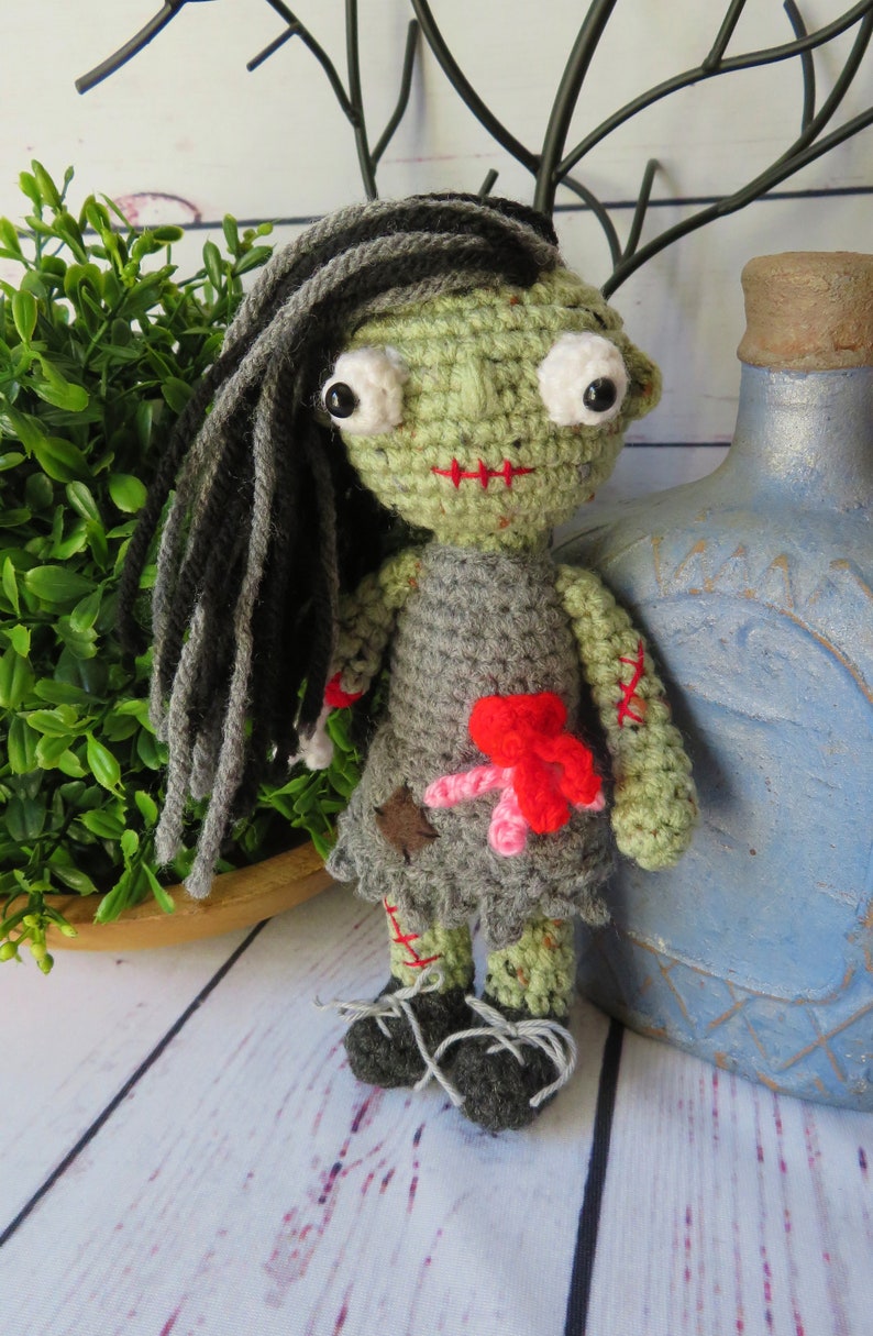 crochet girl zombie with green skin, grey dress and black and grey hair. She has bulging eyes, red scar for mouth, a severed arm with a crochet bone sticking out. She has red and pink intestines coming out of her belly, a red scar one one arm and leg
