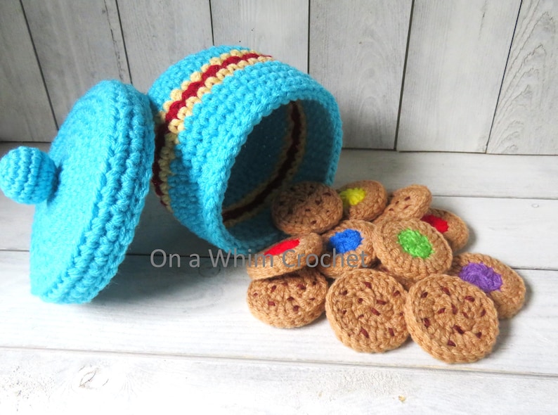 May include: A crocheted cookie jar and cookies. The jar is turquoise with red and yellow stripes. The lid is off, and cookies are spilling out. The cookies are brown with colorful, round decorations. The text "On a Whim Crochet" is visible.