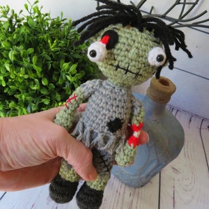 crochet boy zombie shown in hand for size. He is about 8 inches tall. He has green skin, grey clothing, a dislocated eye and a partially severed arm that shows bone and red yarn for blood. He has black messy hair.