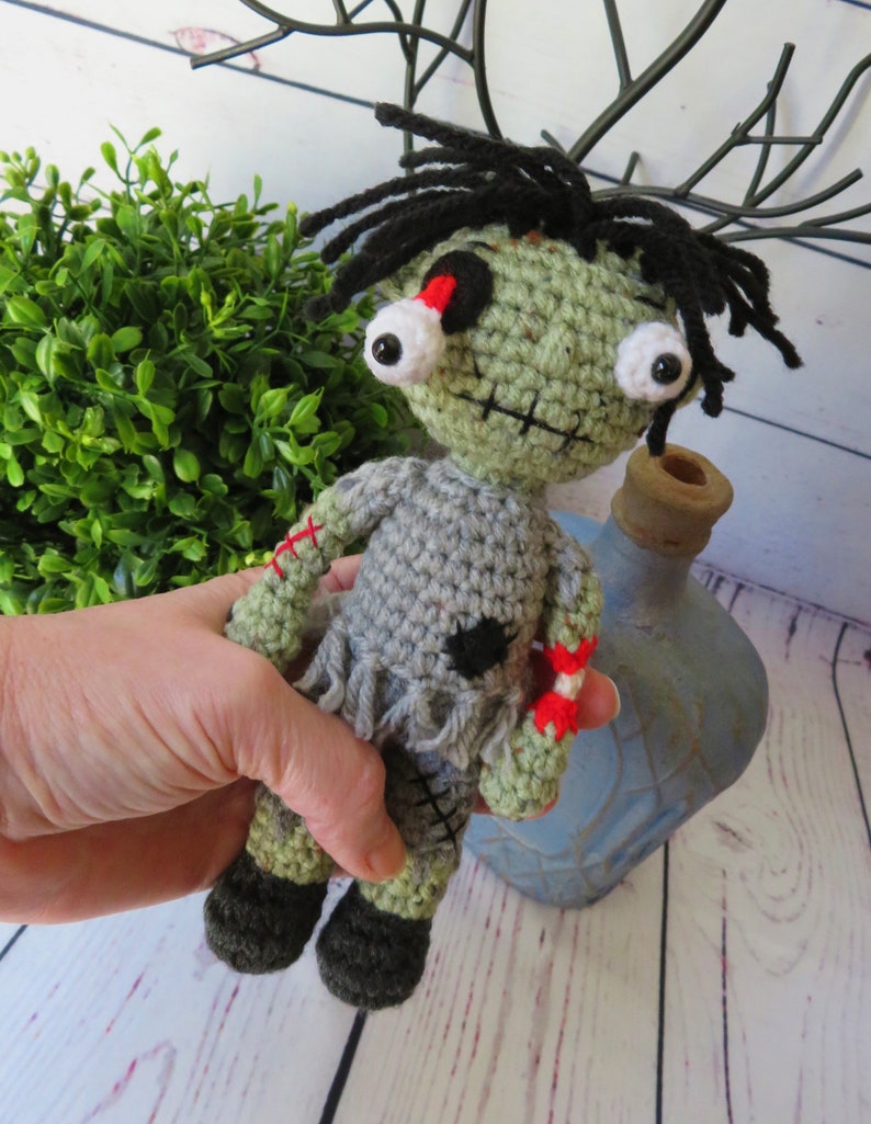 crochet boy zombie shown in hand for size. He is about 8 inches tall. He has green skin, grey clothing, a dislocated eye and a partially severed arm that shows bone and red yarn for blood. He has black messy hair.