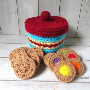 May include: A handmade crochet cookie jar with a red lid and a blue and yellow body. Several crochet cookies with colorful centers are arranged around the jar. The jar and cookies are made of yarn.