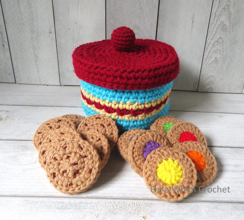 May include: A handmade crochet cookie jar with a red lid and a blue and yellow body. Several crochet cookies with colorful centers are arranged around the jar. The jar and cookies are made of yarn.
