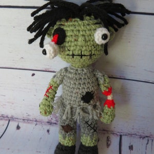 Crochet boy zombie with green skin, grey clothing, a dislocated eye and a partially severed arm that shows a white bone between the upper and lower arm and red yarn for blood. He has black messy hair, an embroidered scar for a mouth.