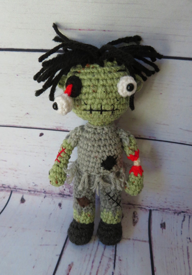 Crochet boy zombie with green skin, grey clothing, a dislocated eye and a partially severed arm that shows a white bone between the upper and lower arm and red yarn for blood. He has black messy hair, an embroidered scar for a mouth.