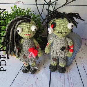 Crochet zombie couple. crochet girl zombie with black and grey hair, green skin, bulging eyes, grey dress, red scars, intestines spilling from belly. Boy zombie with a dislocated eye, grey shirt and pants, red scar dislocated arm with bone and blood.