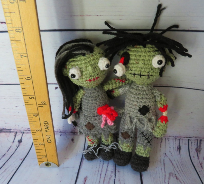 Crochet zombie couple shown next to a ruler to show they are about 7 and a half to 8 inches tall. The zombies have green skin, grey tattered clothing and black messy hair. they have bulging eyes and dislocated arms.
