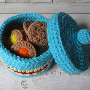 May include: A handmade, open turquoise crochet basket filled with crochet cookies. The basket has a red and yellow trim. The lid is turquoise with a small ball on top. The cookies are brown, yellow, and orange.