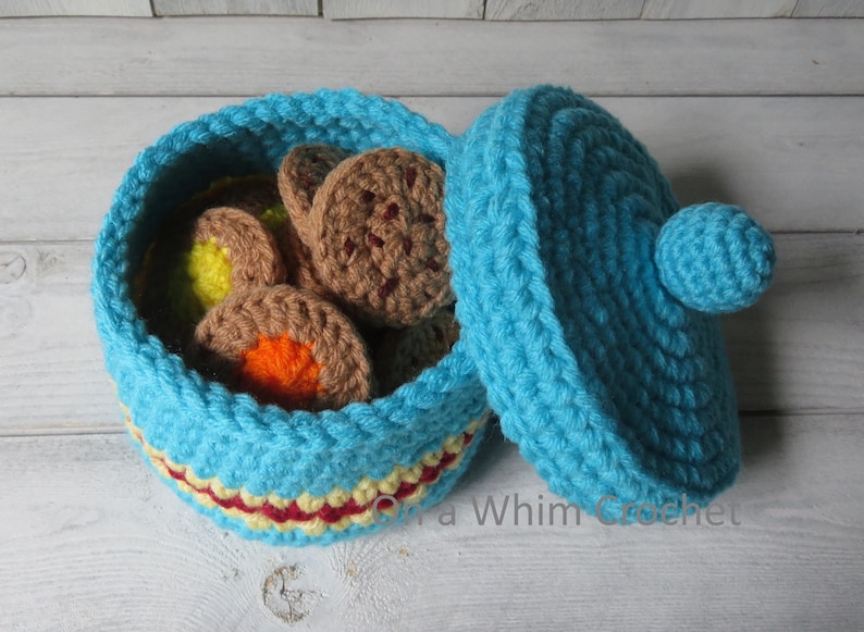 May include: A handmade, open turquoise crochet basket filled with crochet cookies. The basket has a red and yellow trim. The lid is turquoise with a small ball on top. The cookies are brown, yellow, and orange.