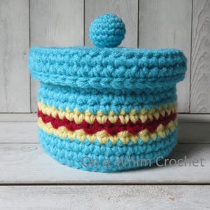 May include: A crocheted container with a lid, in shades of turquoise, red, and yellow. The container has a cylindrical shape with a decorative band of red and yellow. The lid is topped with a small turquoise ball. The text "On a Whim Crochet" is visible.