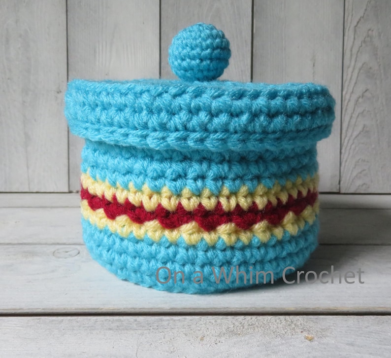 May include: A crocheted container with a lid, in shades of turquoise, red, and yellow. The container has a cylindrical shape with a decorative band of red and yellow. The lid is topped with a small turquoise ball. The text "On a Whim Crochet" is visible.