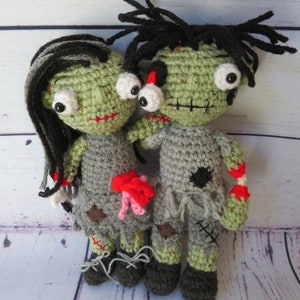 Crochet zombie couple. crochet girl zombie with black and grey hair, green skin, bulging eyes, grey dress, red scars, intestines spilling from belly. Boy zombie with a dislocated eye, grey shirt and pants, red scar dislocated arm with bone and blood.