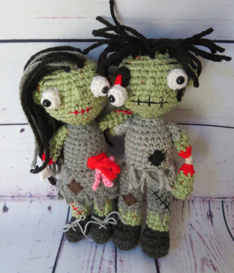 Crochet zombie couple. crochet girl zombie with black and grey hair, green skin, bulging eyes, grey dress, red scars, intestines spilling from belly. Boy zombie with a dislocated eye, grey shirt and pants, red scar dislocated arm with bone and blood.