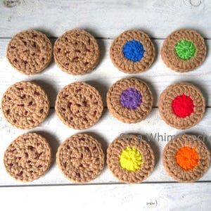 May include: A collection of twelve handmade crochet coasters, resembling cookies. The coasters are primarily tan with darker brown accents, and some feature colorful centers in blue, green, purple, red, yellow, and orange. The coasters are arranged on a light wood surface.