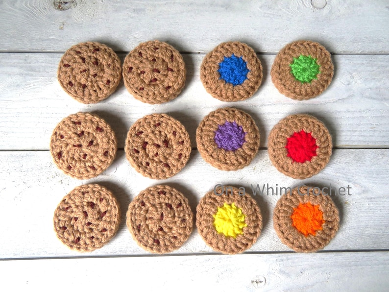 May include: A collection of twelve handmade crochet coasters, resembling cookies. The coasters are primarily tan with darker brown accents, and some feature colorful centers in blue, green, purple, red, yellow, and orange. The coasters are arranged on a light wood surface.