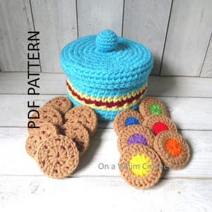 May include: A crocheted blue container with a lid and a red and yellow stripe. Several crocheted cookies are arranged around the container. Some cookies have colorful centers. The text "PDF PATTERN" is on the left side of the image.