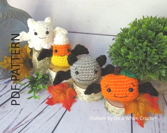 May include: A collection of handmade crochet Halloween decorations, including a white ghost, a candy corn, a gray bat, and an orange pumpkin. Each character has a smiling face and black wings. The text "PDF PATTERN" is visible on the left side of the image.