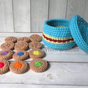 May include: A crocheted cookie jar and cookies. The jar is light blue with yellow and red stripes. The lid is light blue with a small ball on top. The cookies are brown with colorful centers. The text "Whim Crochet" is visible.
